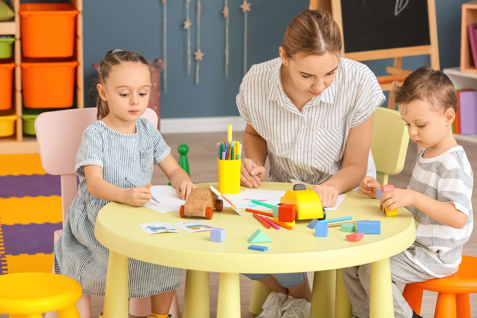 A childminder sits with two youngsters as they do papercrafts.